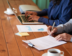 Two people at a wooden table drafting a research proposal: one types on a laptop while the other makes notes on printed charts and forms, with coffee cups nearby