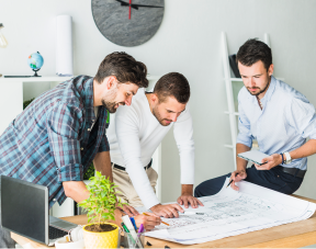Three colleagues gathered around a large desk studying project plans and blueprints, one pointing at the papers while the others take notes