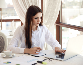 A focused young woman sitting at a desk by a window, typing notes on a laptop with a notebook and pen nearby, appearing to write a reflective journal